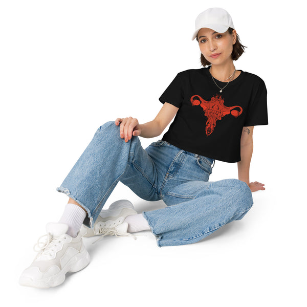 Woman wearing a black crop top with red death metal uterus design, paired with jeans and white sneakers, showcasing a relaxed, witchy aesthetic.